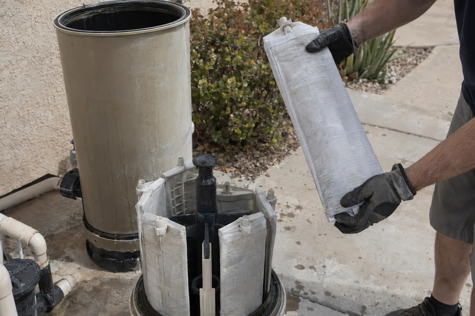 A disassembled diatomaceous earth pool filter with the tank open and grid assembly removed, a technician's gloved hands inspecting one filter grid on a concrete patio in a Southern California backyard