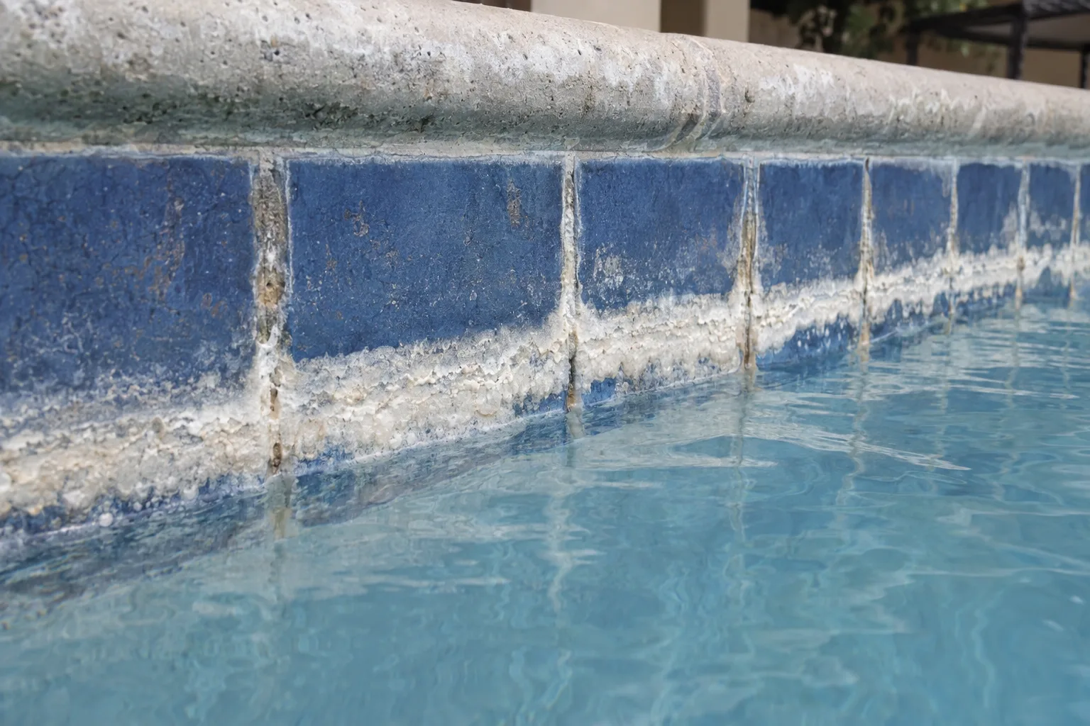 Close-up of thick white calcium scale deposits crusted along the waterline tiles of a swimming pool, with clear blue water visible just below the mineral buildup