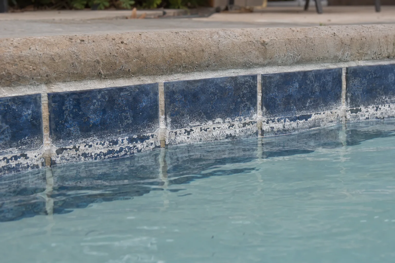 Heavy white calcium scale deposits crusted along the blue waterline tile and grout of a residential swimming pool in bright Southern California sunlight